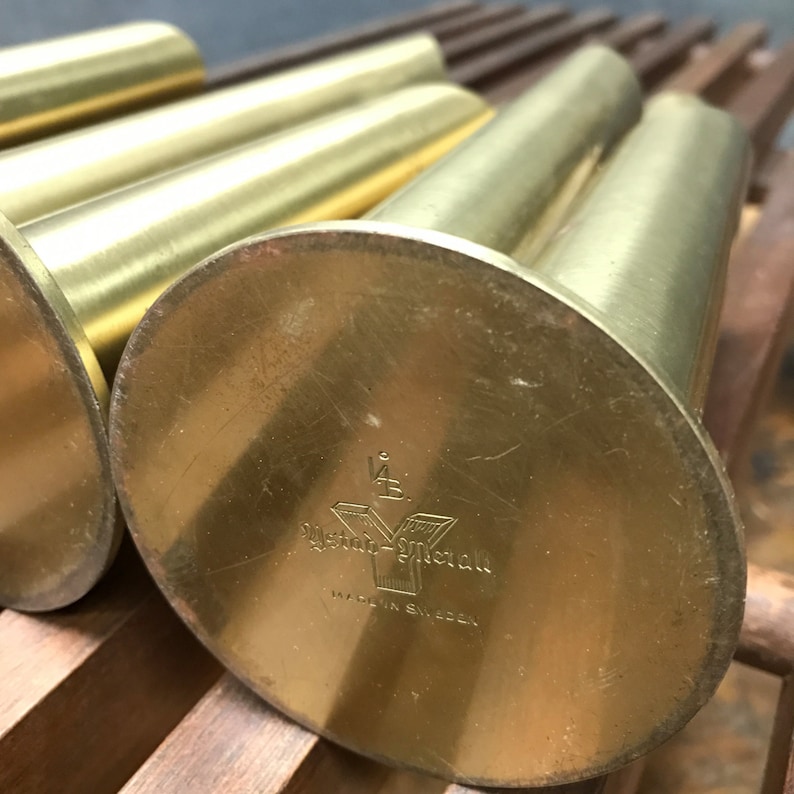 May include: Close-up of the bottom of a set of four gold metal candle holders. The bottom of each holder has a circular base with the text "Ystad-Metall Made in Sweden" engraved on it.