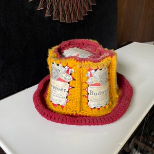 May include: A crocheted hat made from Budweiser beer cans. The hat is yellow and red with a wide brim.