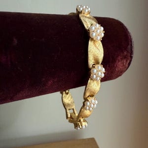 Op de afbeelding: Een goudkleurige armband met een bladeren design, versierd met clusters van kleine, witte parels. De armband wordt gepresenteerd op een donkerrood fluwelen oppervlak, wat de elegante en vintage stijl benadrukt. De sluiting is zichtbaar, wat duidt op een veilige sluiting.