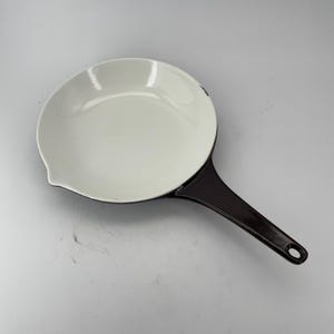May include: A white enamel frying pan with a dark brown handle. The pan has a rounded shape with a pouring spout. The handle has a hole at the end for hanging. The pan is designed for cooking.