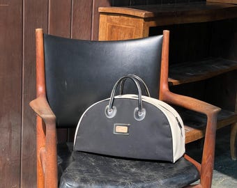 Pode incluir: Uma mala de mão preta e creme com um design semicircular, alças pretas e uma pequena etiqueta retangular. A mala está colocada numa cadeira vintage de couro preto com apoios de braços de madeira e uma estrutura de madeira.