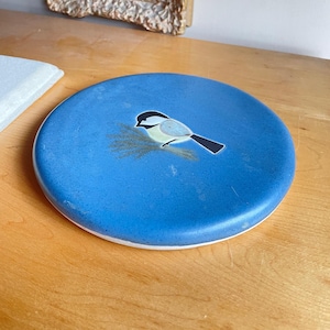 Op de afbeelding: Een blauw keramiek bord met een witte rand heeft een geschilderd beeld van een kleine vogel met een zwarte kop en gele borst. De vogel zit op een tak van twijgen.