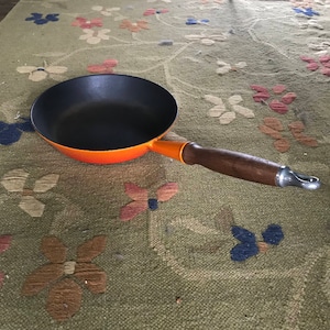 Puede incluir: Sartén de hierro fundido naranja con mango de madera. La sartén tiene un interior negro y un exterior naranja oscuro.