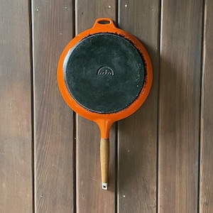 May include: An orange cast iron skillet with a wooden handle. The skillet has a black cooking surface and a loop for hanging. The handle is light brown and attached to the orange skillet. The skillet is hanging on a wooden wall.