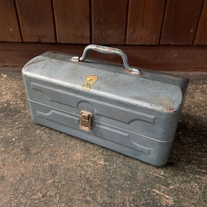 May include: A vintage, light blue metal toolbox with a handle and a latch. The toolbox has a rectangular shape with decorative indentations on the sides. A small sticker is visible on the front.