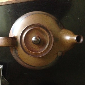 Può includere: Una teiera in ceramica marrone con un coperchio e un manico. La teiera ha una finitura liscia e smaltata.