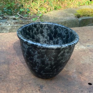 May include: A round, bowl-shaped planter featuring a dark black base color with a mottled, speckled pattern in shades of light gray and charcoal. The vessel has a smooth, rounded rim and a deep, open interior, showcasing a consistent finish.