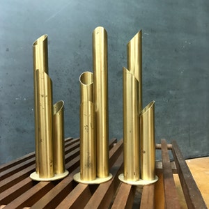 May include: Three gold metal vases with a cylindrical shape and multiple openings. The vases are arranged in a row on a wooden surface.
