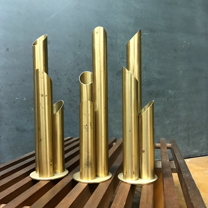 May include: Three gold metal vases with a cylindrical shape and multiple openings. The vases are arranged in a row on a wooden surface.