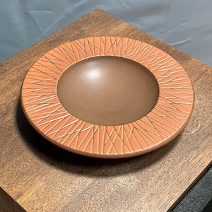 May include: A round ceramic bowl with an orange rim and a brown interior. The rim has a repeating pattern of white lines. The bowl sits on a wooden surface.