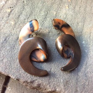 Può includere: Un paio di orecchini a spirale di colore marrone e nero. Gli orecchini sono realizzati in un materiale lucido e liscio.