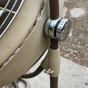 Peut inclure: Un ventilateur oscillant vintage en m&eacute;tal blanc avec une base en m&eacute;tal brun et un bouton de r&eacute;glage chrom&eacute;. Le ventilateur a une cage circulaire en fil m&eacute;tallique et une grande pale ronde.