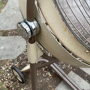 Peut inclure: Un ventilateur oscillant vintage en m&eacute;tal brun avec une grille de protection en plastique blanc et un bouton chrom&eacute;. Le ventilateur est sur un pied avec des roulettes.