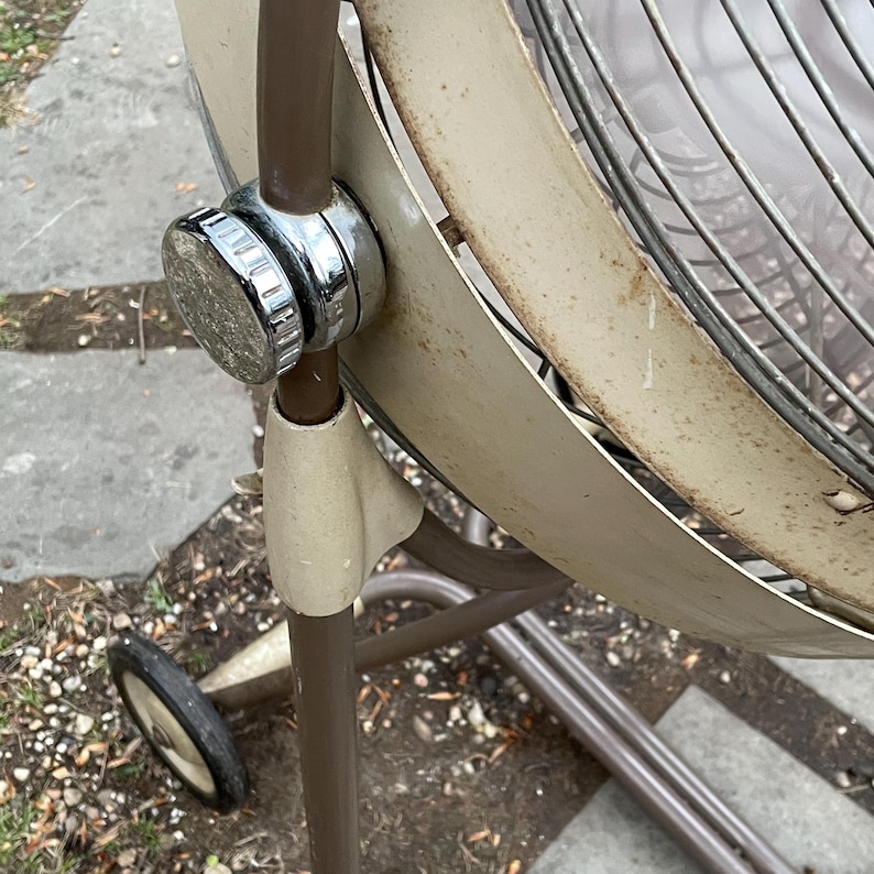 Peut inclure: Un ventilateur oscillant vintage en m&eacute;tal brun avec une grille de protection en plastique blanc et un bouton chrom&eacute;. Le ventilateur est sur un pied avec des roulettes.