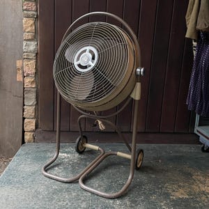 Peut inclure: Un grand ventilateur vintage en m&eacute;tal avec une finition brune et argent&eacute;e. Le ventilateur est sur un support roulant avec deux roues et une base en m&eacute;tal. Le ventilateur est devant un mur en bois.