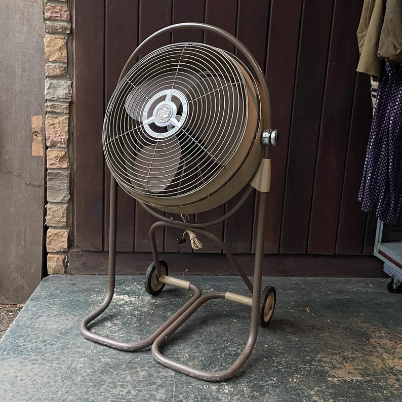 Peut inclure: Un grand ventilateur vintage en m&eacute;tal avec une finition brune et argent&eacute;e. Le ventilateur est sur un support roulant avec deux roues et une base en m&eacute;tal. Le ventilateur est devant un mur en bois.