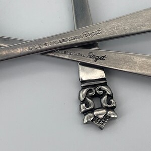 May include: Close-up of two silver spoons with the words "Stainless Japan  Rogert" engraved on the back of the spoons. The spoons have an ornate design at the end.