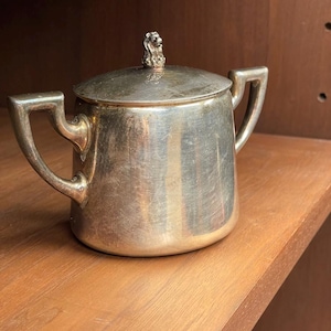 May include: A silver-toned metal sugar bowl with two angular handles and a matching lid. The lid features a small, sculpted lion finial at the center. The bowl has a smooth, polished surface with visible signs of wear and light tarnishing throughout.
