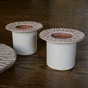 May include: Two white ceramic candle holders with a raised, textured design around the top rim. The design is in a light brown color. The candle holders are on a wooden surface.