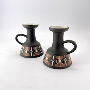 May include: Two black ceramic candle holders with a brown and white geometric pattern. Each holder has a handle and a flared base. The interior of the holders is white.