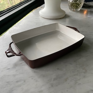Puede incluir: Un molde para hornear rectangular con un interior blanco y un exterior marrón oscuro. El molde tiene dos asas a los lados.