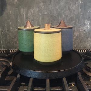 Puede incluir: Tres tarros de cerámica con tapa de diferentes colores. Los tarros son verde, amarillo y azul. Los tarros están sobre un trípode de hierro fundido negro.