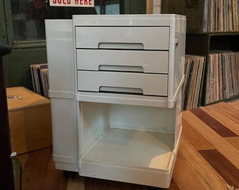 May include: A white, three-drawer cabinet with an open bottom shelf. The cabinet has a rectangular shape and is mounted on small wheels. The drawers have horizontal handles. The sign in the background reads "SOLD HERE".