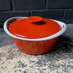 Könnte beinhalten: Orangefarbene und weiße Keramik-Auflaufform mit Deckel. Die Form hat zwei Griffe an den Seiten und einen Knopf auf dem Deckel.