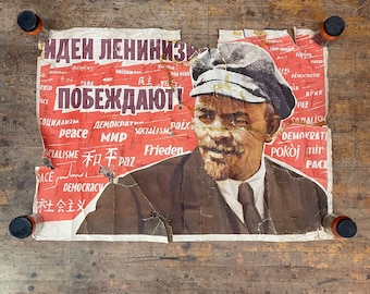 May include: A vintage, torn poster featuring a portrait of a man in a cap, with text in red and other colors. The poster is on a wooden surface, with four small bottles with black caps at each corner.