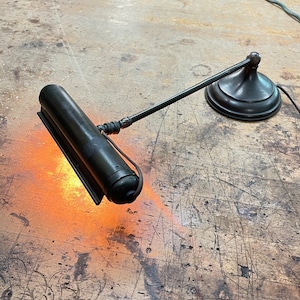 Puede incluir: Una lámpara de escritorio de metal negro con un brazo largo y ajustable y una pantalla cilíndrica. La lámpara está encendida y proyecta una luz cálida y naranja.