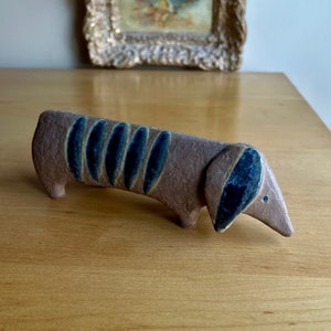 Może przedstawiać: Ceramiczna figurka psa rasy dachshund z brązową i niebieską glazurą. Figurka stoi na drewnianej powierzchni.