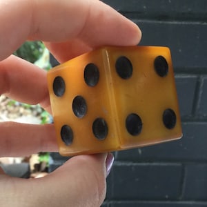 Op de afbeelding: Een grote, amberkleurige dobbelsteen met zwarte stippen. De dobbelsteen wordt in een hand gehouden, wat de grootte en de helderheid van het materiaal laat zien. De stippen zijn gelijkmatig verdeeld, wat duidt op een standaard zeszijdige dobbelsteen. Geschikt voor spellen en verzamelobjecten.
