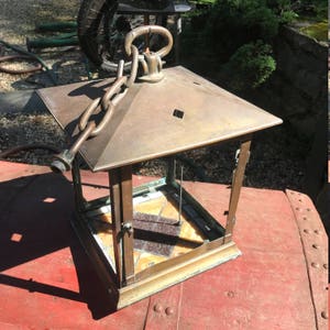 May include: An antique brass lantern with a square, pyramidal top and a chain for hanging. The lantern has glass panels and a decorative stained glass base. The metal has a weathered patina, suggesting age and use. The lantern is sitting on a red surface.