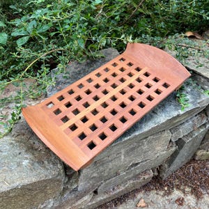 May include: A rectangular wooden tray with a grid of square cutouts. The tray has curved edges and is made of light brown wood. It is resting on a stone wall with green foliage in the background.