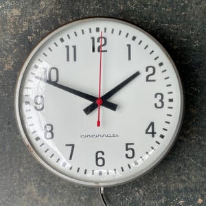 May include: A large, round, white clock face with black hands and black numbers. The clock is in a silver metal frame and has the word "Cincinnati" printed on the face.