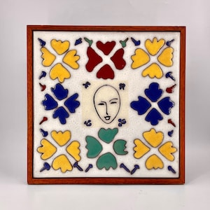 May include: A framed ceramic tile with a white background and a stylized face in the center. The tile is decorated with yellow, red, blue, and green floral designs.