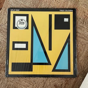 May include: A vinyl record album titled "True Colours" by Split Enz. The album cover features a yellow background with black and light blue geometric shapes. The text "SPLIT ENZ" and "TRUE COLOURS" are visible. A "Value Priced" sticker is on the cover.