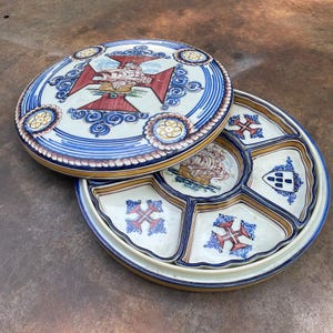 May include: A round ceramic serving dish with a matching lid, featuring a central hand-painted sailing ship, red crosses, and blue scrollwork. The base is divided into six wedge-shaped compartments, each decorated with red crosses or blue shields.