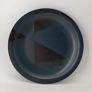 Può includere: Un piatto rotondo blu scuro con un design geometrico. Il piatto presenta una serie di triangoli marroni scuri disposti sulla superficie. Il piatto ha un bordo leggermente rialzato e una finitura opaca. Questo piatto è adatto per servire cibo, circa 25 cm di diametro.