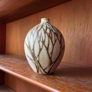 Puede incluir: Un pequeño jarrón de cerámica blanca con un diseño de ramas de árbol marrones. El jarrón es redondo y tiene una abertura estrecha en la parte superior.