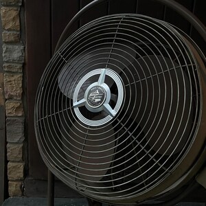 Peut inclure: Un ventilateur vintage Emerson Electric avec une cage en m&eacute;tal noir et une &eacute;tiquette argent&eacute;e qui indique "Roll About Oscillator".