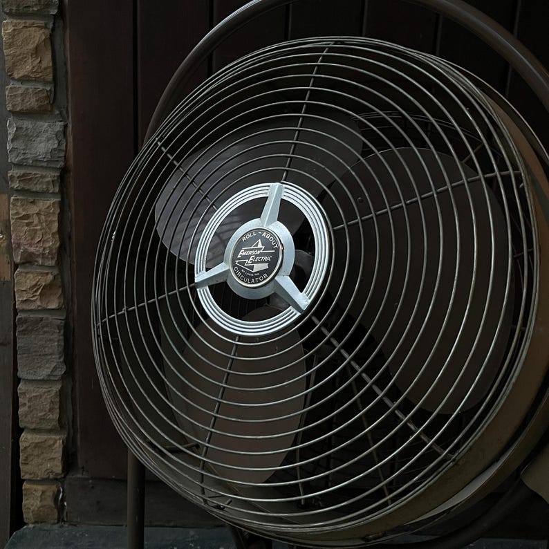 Peut inclure: Un ventilateur vintage Emerson Electric avec une cage en m&eacute;tal noir et une &eacute;tiquette argent&eacute;e qui indique "Roll About Oscillator".