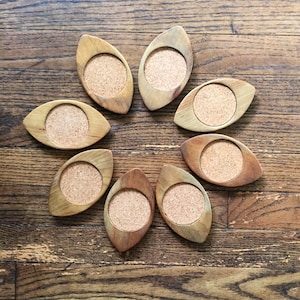 May include: Eight wooden coasters with cork inserts arranged in a circle on a wood surface. The coasters are oval shaped with a light brown wood finish.