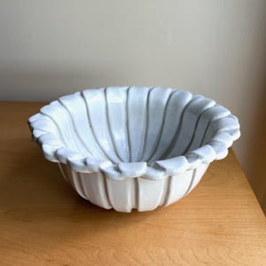 May include: A white ceramic bowl with a scalloped rim and vertical ribbed detailing. The bowl is round and sits on a wooden surface. The bowl's design is reminiscent of a seashell.