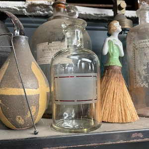 May include: A clear glass bottle with a red rectangular label. The bottle has a glass stopper and is surrounded by other vintage bottles and objects. The background is a shelf with other antique items.