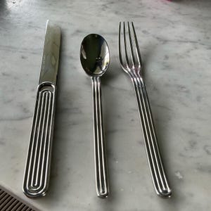 May include: A set of three silver-toned utensils, including a knife, fork, and spoon. Each utensil has a unique, grooved design.