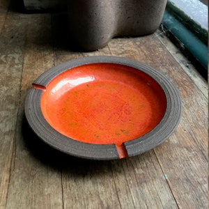 May include: A round orange ceramic ashtray with a dark brown rim. The ashtray has a slightly raised edge and a small indentation on each side.