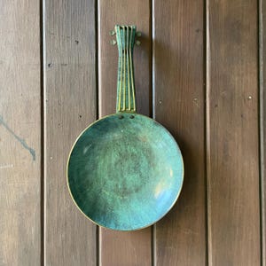 May include: A vintage-style, round, green-patinaed metal pan with a long handle. The handle has vertical gold-colored stripes. The pan is mounted on a wooden surface. The pan's circular bowl shape is clearly visible.