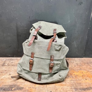 May include: A green canvas backpack with brown leather straps and buckles. The backpack has a flap closure and a large main compartment.
