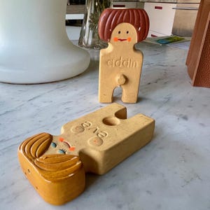 May include: Two ceramic figurines of people with painted faces and hair. The figurines are nude and have a hole in the center of their bodies. The figurine on the left is lying down and the figurine on the right is standing up. The figurine on the right has the word "cilddin" written on its torso.